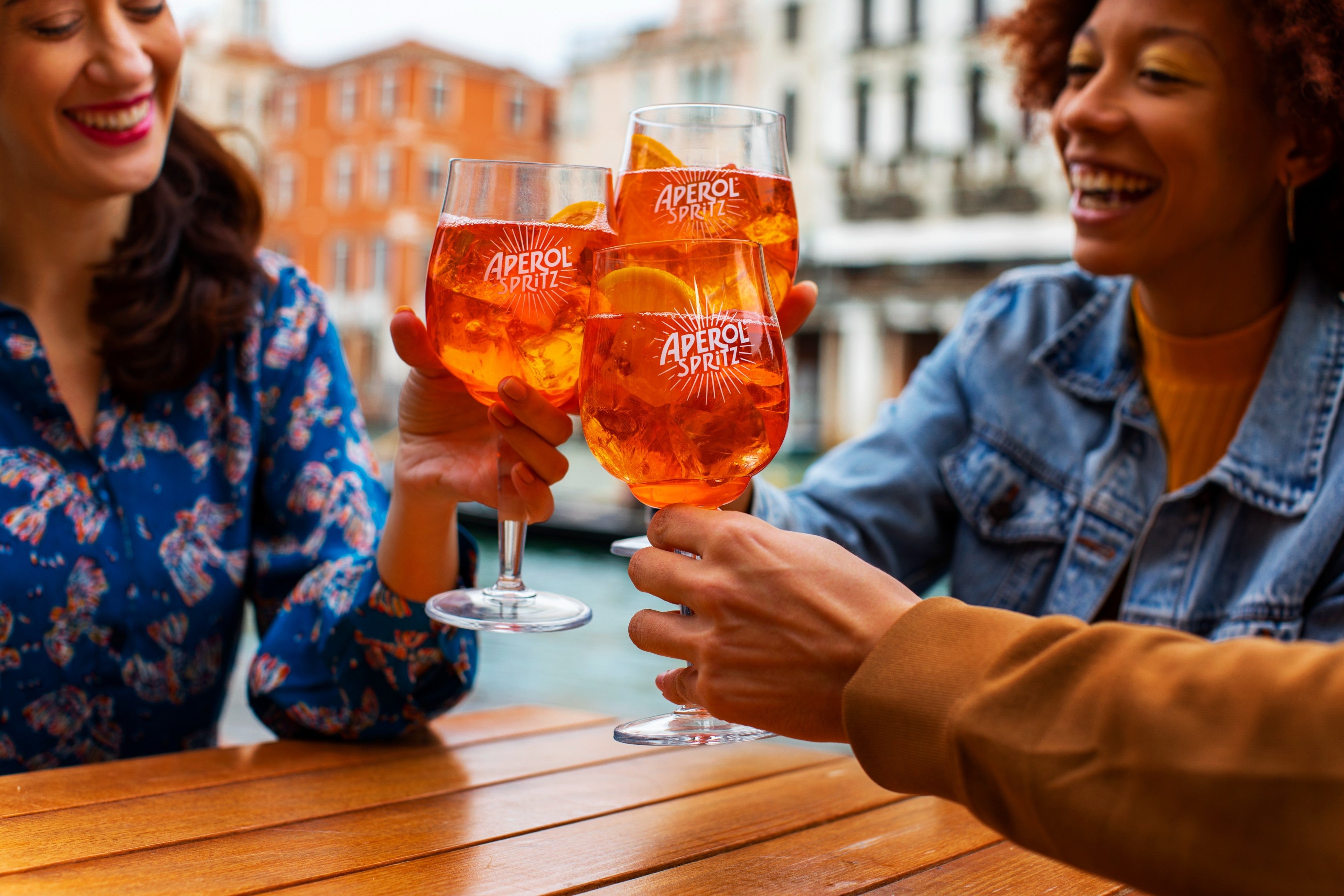 Three friends cheerfully clink their Aperol Spritz glasses while seated at a wooden outdoor table in a vibrant European city setting, with colorful buildings and a canal in the background.